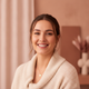 A smiling young woman, headshot portrait, looking confidently at the viewer. Background is soft and blurred in warm tones.
